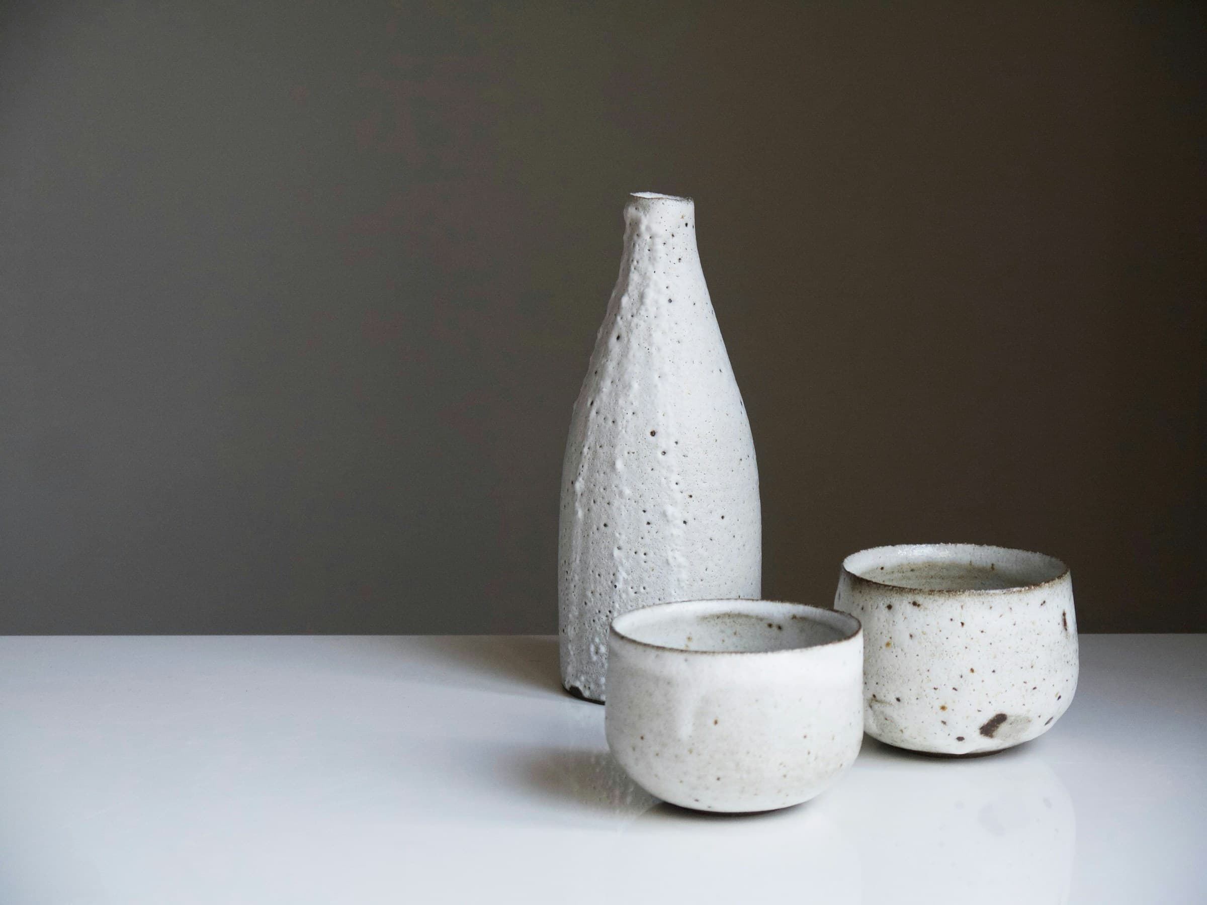 A glazed stoneware bottle and two speckled stoneware cups on a white surface against a warm grey wall.